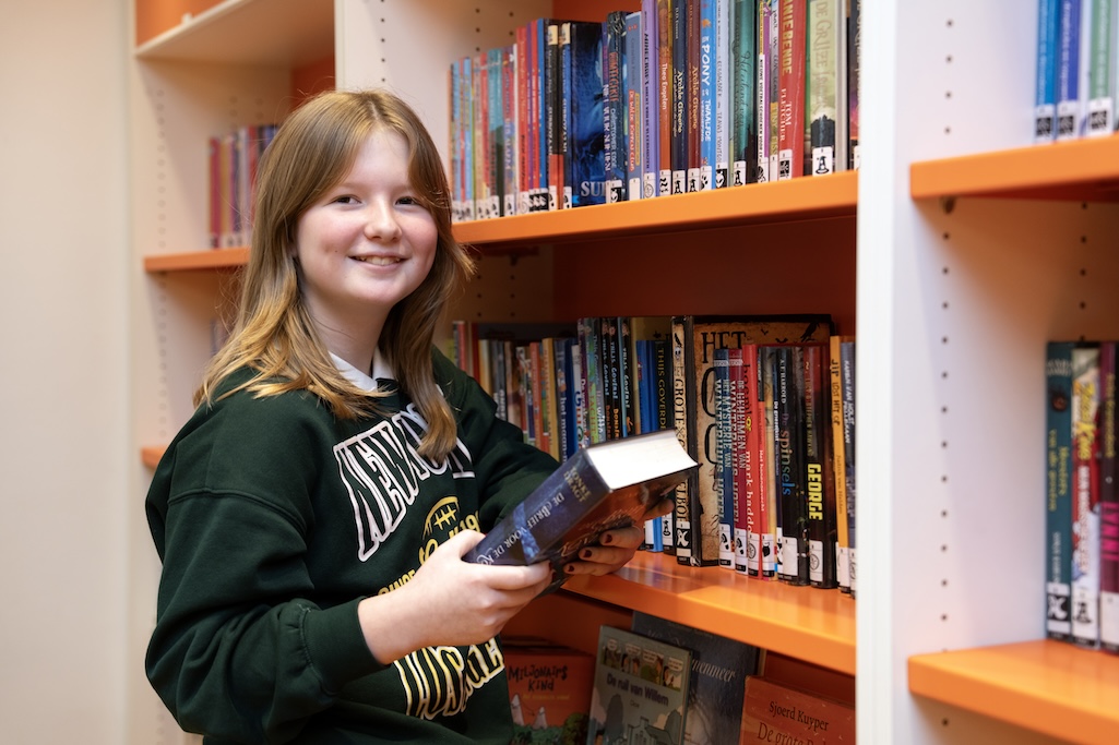 Meisje kijkt lachend met boek in haar hand de camera in, achter haar een oranje boekenkast van de Bibliotheek